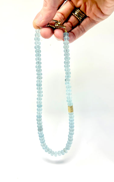 A beaded necklace made of light blue faceted beads with gold-toned metal hardware, held by a hand against a plain white background.