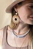 Woman wearing a large sunflower-shaped earring and multiple necklaces with a neutral background