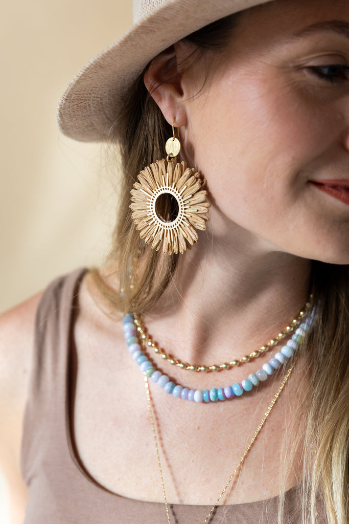 Woman wearing a large sunflower-shaped earring and multiple necklaces with a neutral background