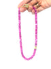 A pink beaded necklace with a gold toggle clasp and a small gold spacer bead, held by a hand against a white background.